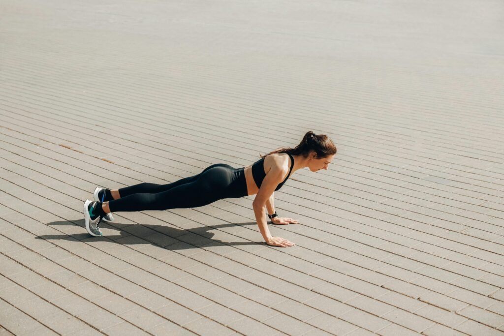 A woman in activewear performs a plank exercise outdoors on a sunny day.